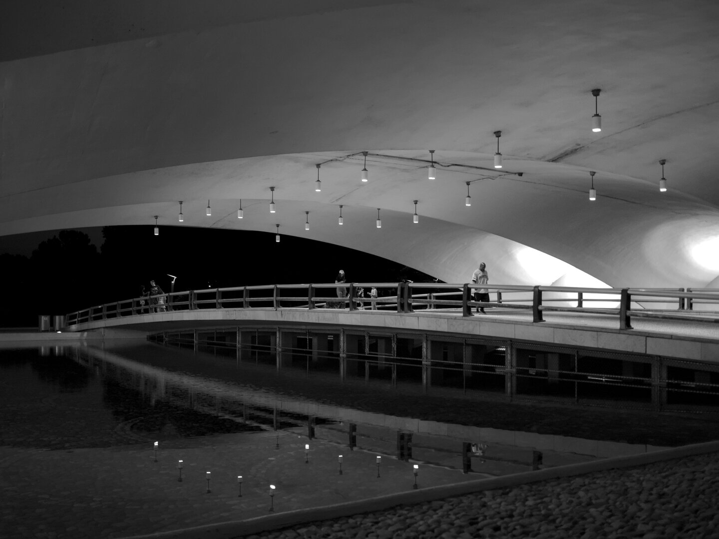 Black and white photo showing a sleek modern flat vaulted tunnel lit from the sides. There's a little pedestrian bridge over a pond that reflects the people on the bridge.