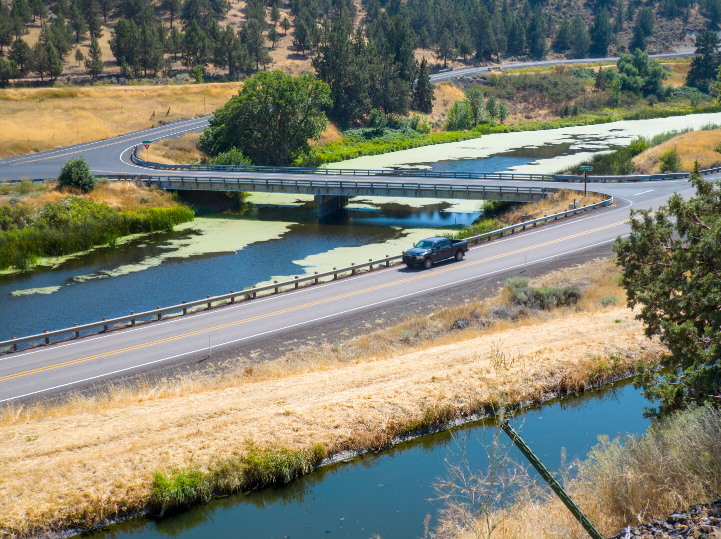Looking down at the canal and the river, showing 2 lane roads on both sides of the river. There's a road bridge across the river. A black pickup truck is traveling on the road on my side of the river.