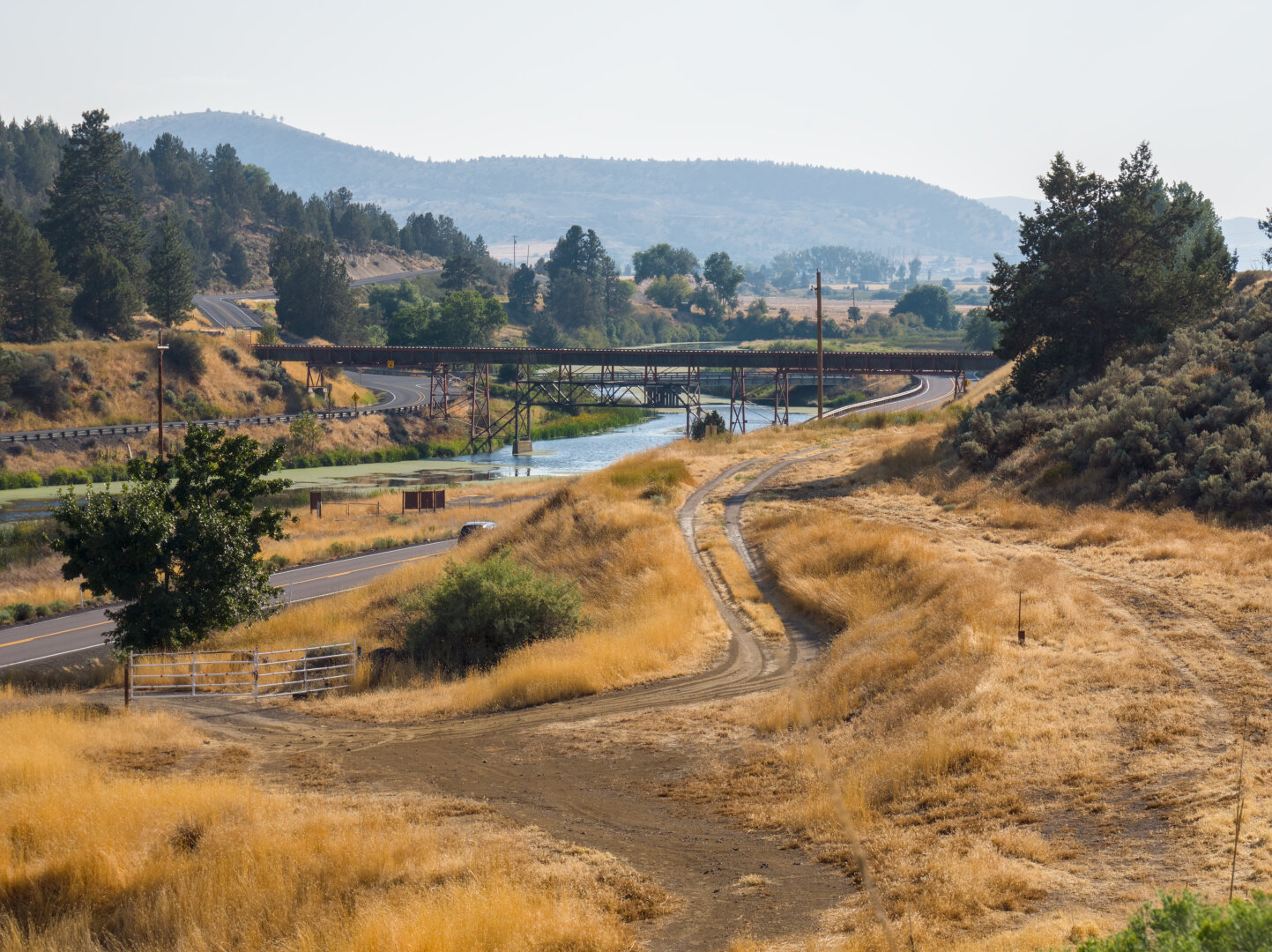 Looking down a hill covered by arid yellow grass with a gravel double track. Beneath the hill is a paved 2 lane road and a river. A small railroad bridge crosses this river. On the other side of the river is a greener hill.