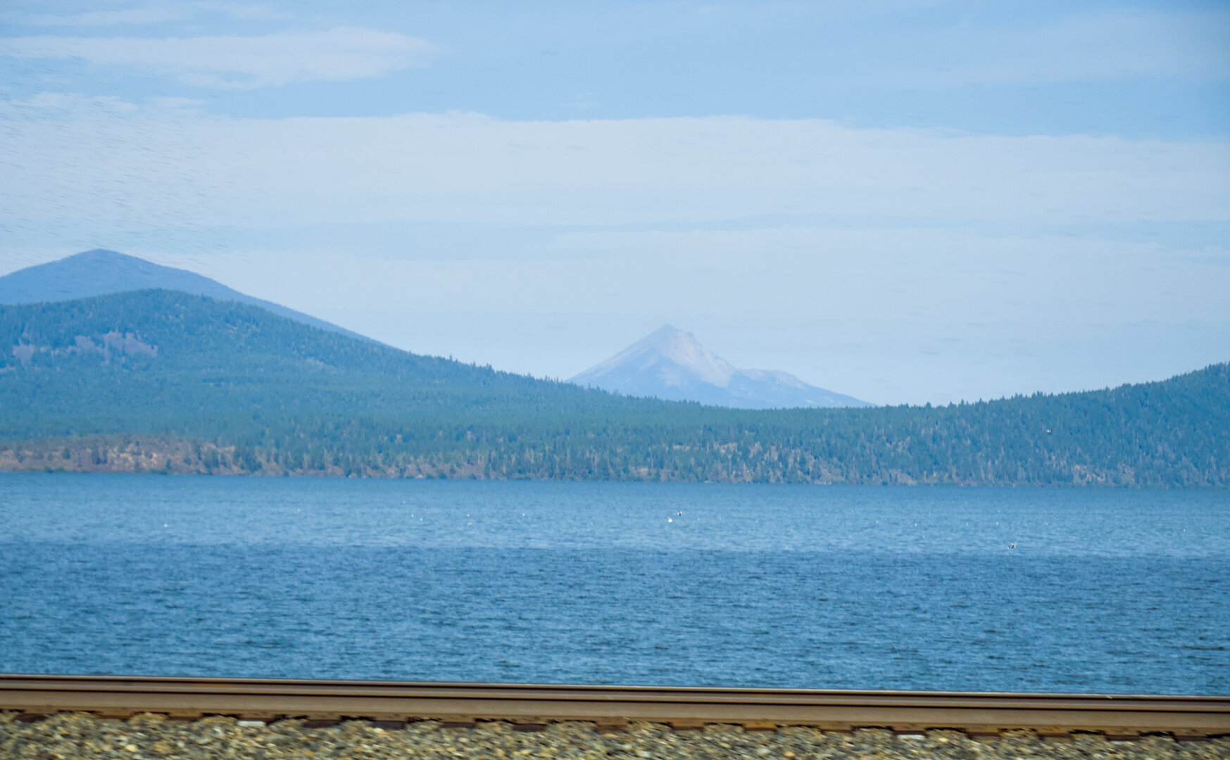 A nearer mountain by a vast lake, and a pointy volcano Mt McLoughlin in the distance. In the foreground is a railroad track and ballast