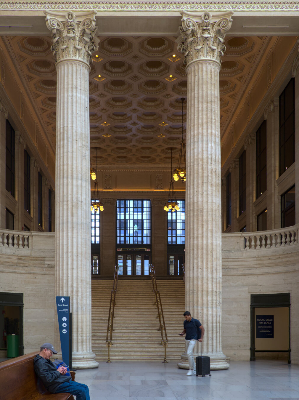 Grand Corinthian neo-classical columns support a grand  hallway leading outside to the streets, up a flight of stairs. The column dwarfs the people nearby. A person in dark gray shirt was sitting on the wooden bench, and another person was walking with a small black suitcase.