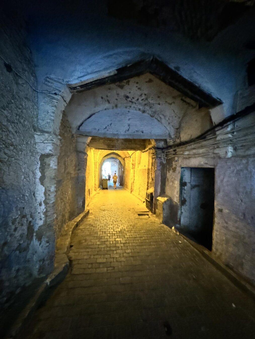 a dark stone street. further on you can see a fragment lit by lanterns and the sun