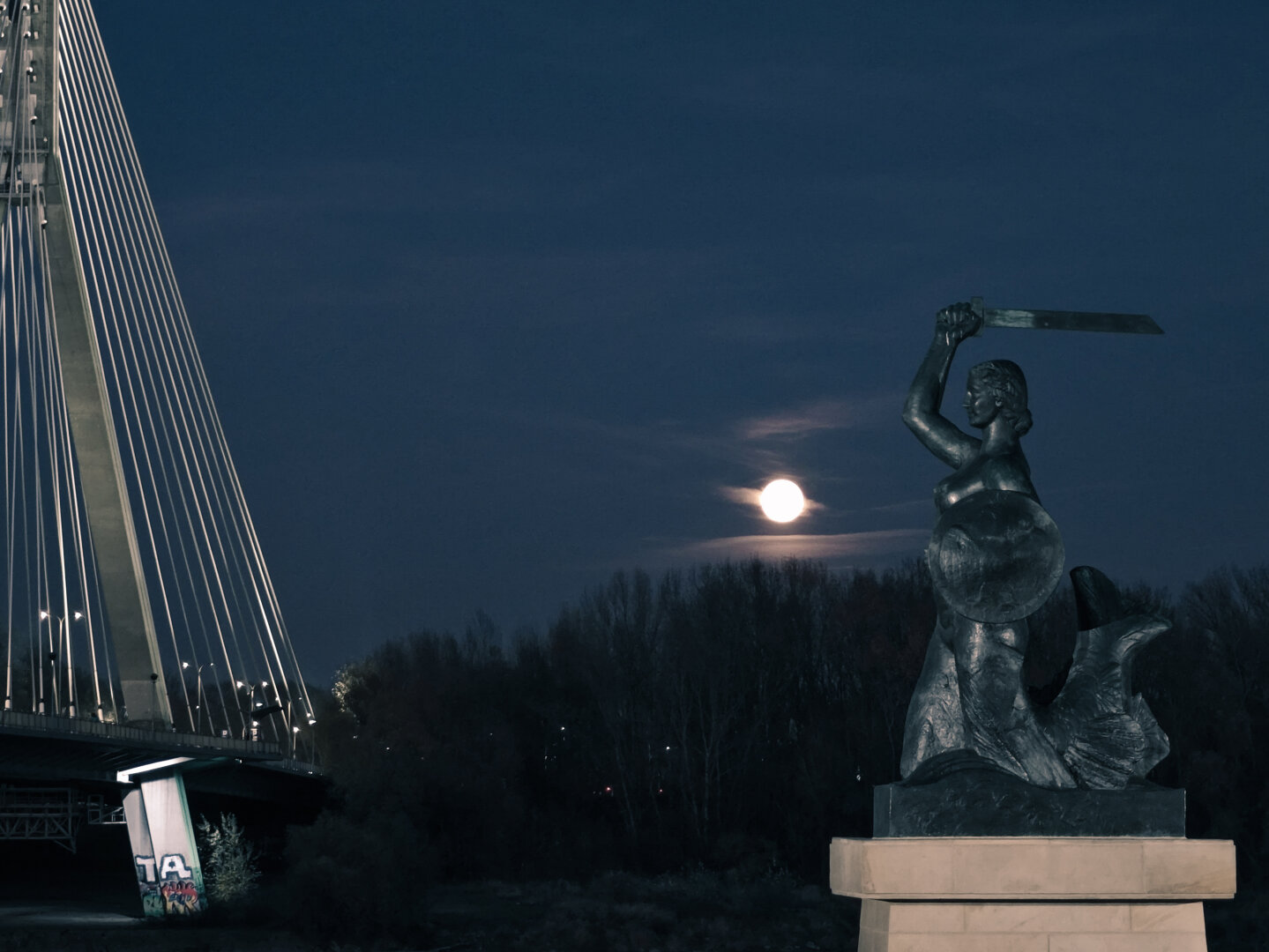(PL): Nocne zdjęcie warszawskiej Syrenki nad Wisłą, z uniesionym mieczem i tarczą, na tle pełni księżyca. Po lewej stronie widać fragment mostu Świętokrzyskiego z oświetlonymi linami i pylonem. Scenę rozświetla delikatne, chłodne światło księżyca

(EN): Nighttime photo of the Warsaw Mermaid statue by the Vistula River, holding a raised sword and shield against the backdrop of a full moon. On the left side, part of the illuminated Świętokrzyski Bridge with its cables and pylon is visible. The scene is softly lit by the cool moonlight