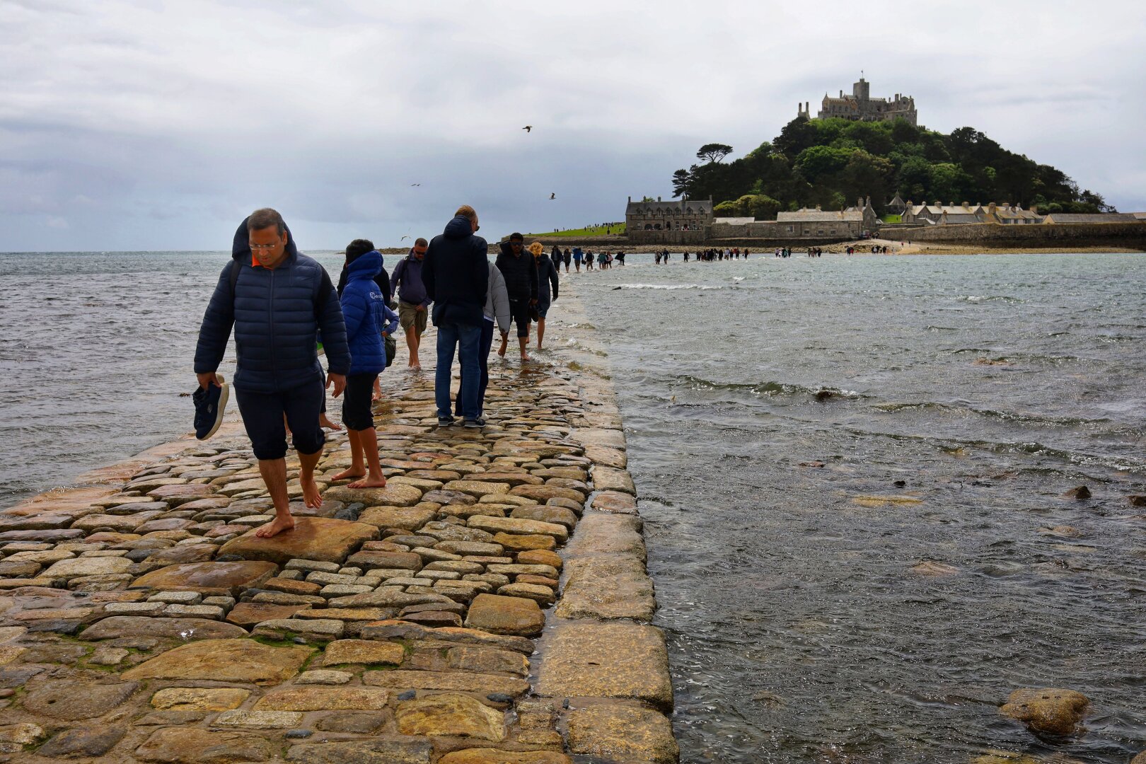 People walking on a causeway. The sea is partially covering the path. People are wading through the water. A small island with a castle is visible at the other end of the causeway. People walking on a causeway. The sea is partially covering the path. People are wading through the water. A small island with a castle is visible at the other end of the causeway.
