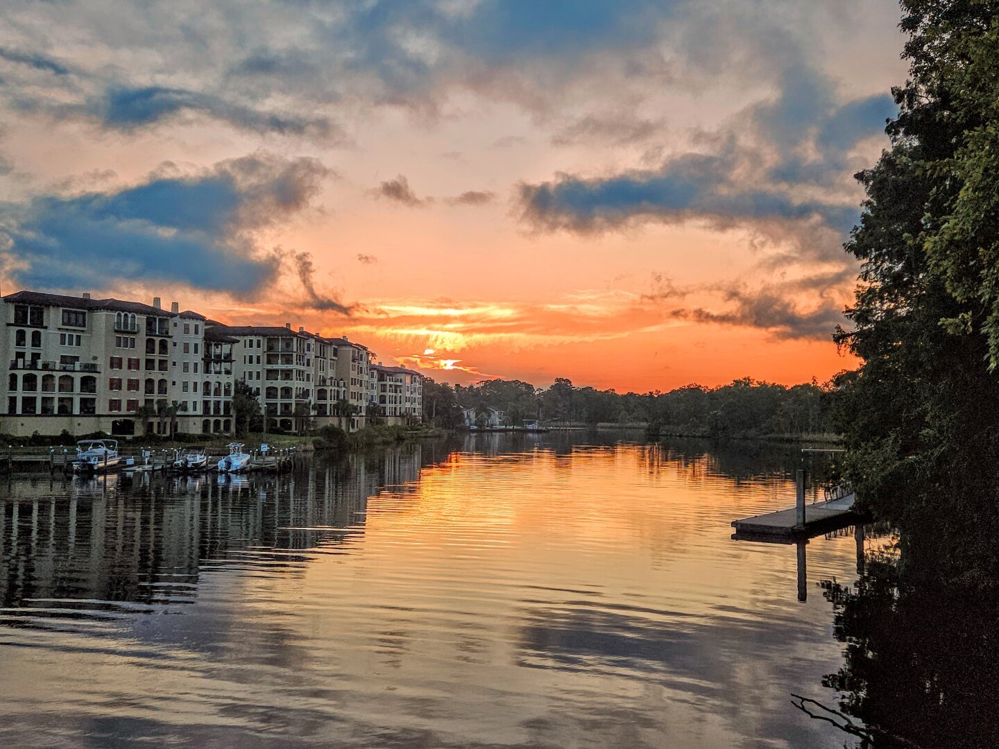 Firey sunrise over a quiet waterway with large luxurious buildings on the far shoreline and tropical swamp foliage in the shadows of the near shoreline. Bright warm shades of red and orange rise from the horizon sending a glow across the morning sky and casting a golden reflection upon the water below.