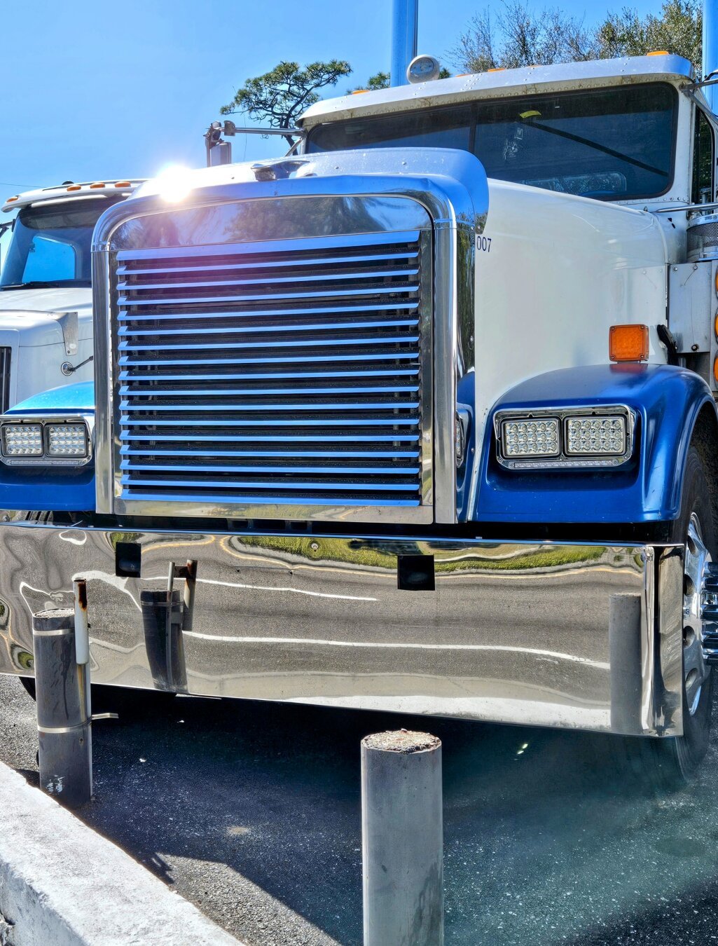 Standing in front of an enormous semi-truck cab on a bright sunny day. The sun's light bounces off the large silver grill on the cab's front.