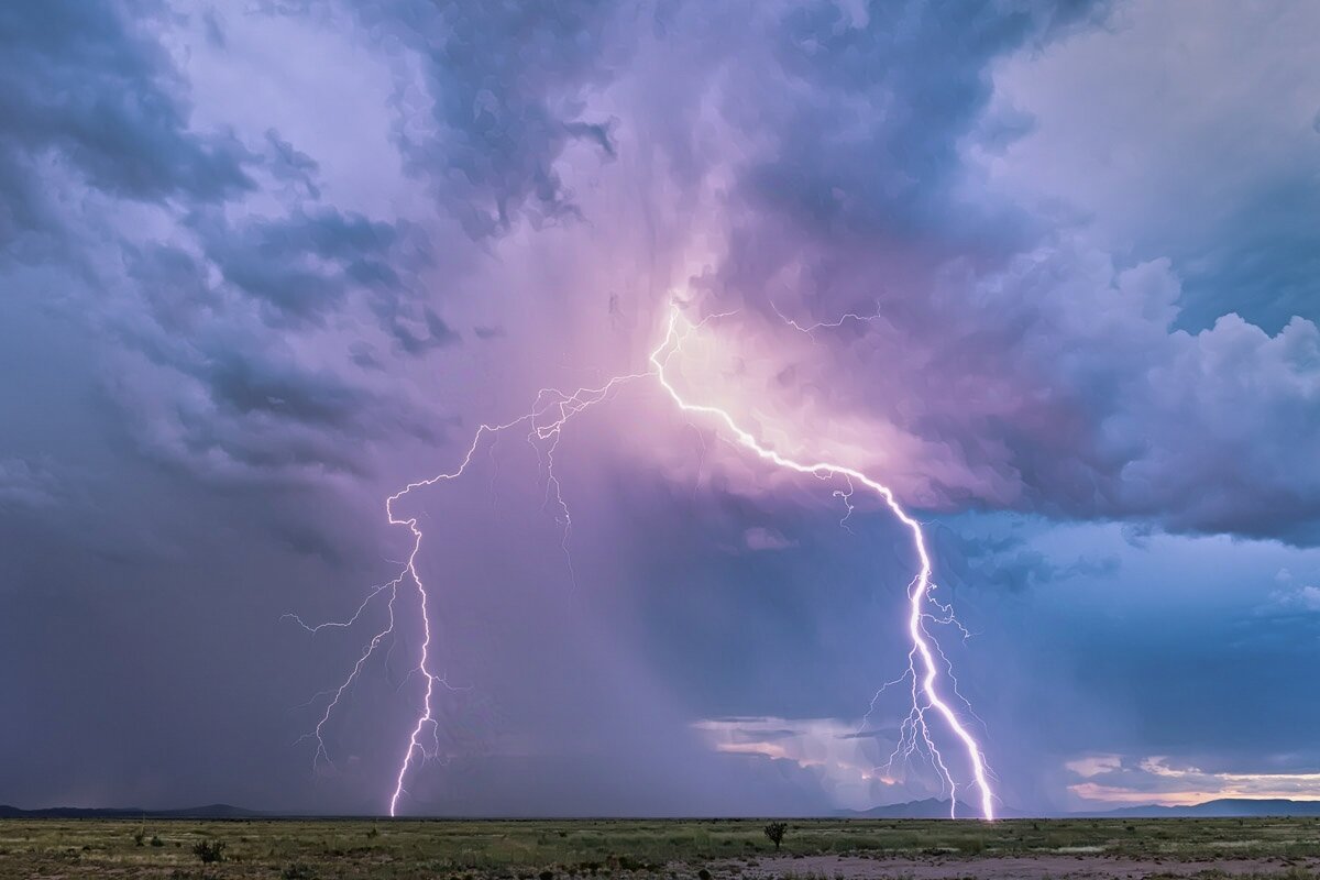 The photograph shows an empty field with low distant hills in the background. It’s late summer, so the field has some green, but there’s also a bare patch of dirt. There is a single small cactus. Just a bit of the field is showing, so the photograph is mostly sky.

The sky is full of dark, ominous clouds, full of blues and purples. Why purple? Because the clouds are lit by twin strikes of lightning, cloud to ground. The bolts emanate from the top center of the photograph, and it’s their light that makes the clouds grow.

These are large bolts, especially the one on the right. They are impressive, and you can feel the surge of electricity in the photograph. There are numerous smaller tendrils branching off the main bolt. You can feel the tension and the drama in this photograph. And yes, the thunder from this strike was epic.

I was obviously close to the lightning, and one could argue far too close. Lightning is deadly and completely unpredictable. If you can see it, it can see you. An