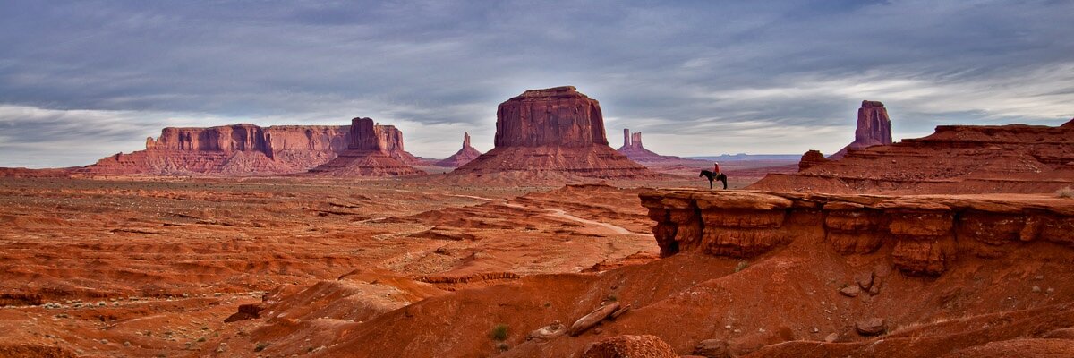 A man on a horse watches over Monument Valley

A man in a red shirt is sitting on a standing horse right of center in this photograph. The man and horse are on a rock outcrop. You might think the man and horse dominate the photograph, but that’s not the case. They’re quite small, and you have to squint to make out any details. The landscape is vast, and they are but a tiny part of it. And that, in turn, truly emphasizes the scale.

Behind them is a large portion of Monument Valley. 1,000 foot buttes look almost small, but they’re not. But the photograph encompasses so much that your sense of scale is impacted. 

The skies are cloudy with some thick clouds. Not the kind that are likely to produce rain, but just thick. The scene is sunlit, though, because there was a break in the clouds that allowed the sun to shine through.