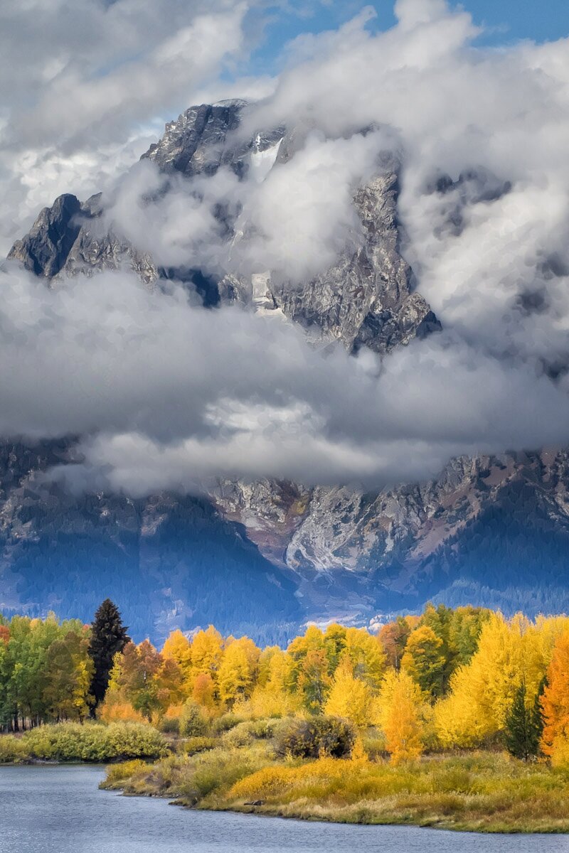 The snake flows past the cloud-wrapped Mount Moran with aspens in full fall color.

This photograph is oriented vertically. The Snake River takes up the very bottom of the photograph. There are a few ripples on it, but overall it is pretty calm. Aspens line the far bank, and they are in full fall color. There’s also a single pine tree there. It’s looking pretty lost in and among the aspens, that’s for sure. 

Above the aspens and taking up the top two thirds of the photograph is Mount Moran. You can’t see it very well because it is cloud-wrapped, but there are a few breaks here and there that let the mountain peak through. You can see, barely, that it has a little snow here and there.

There’s a hint of blue sky in the upper right, but again, it’s mostly clouds.

Overall, it makes a strong impression of the mountain towering over fall.