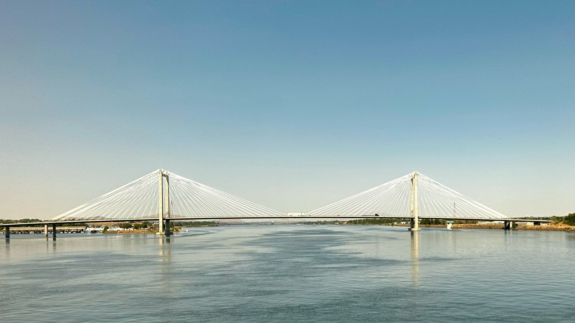 A road bridge with white cables over a large river. The sky fades from blue on the top to whiter toward the bottom due to haze from smoke and pollution, but it is not oppressively smoky.