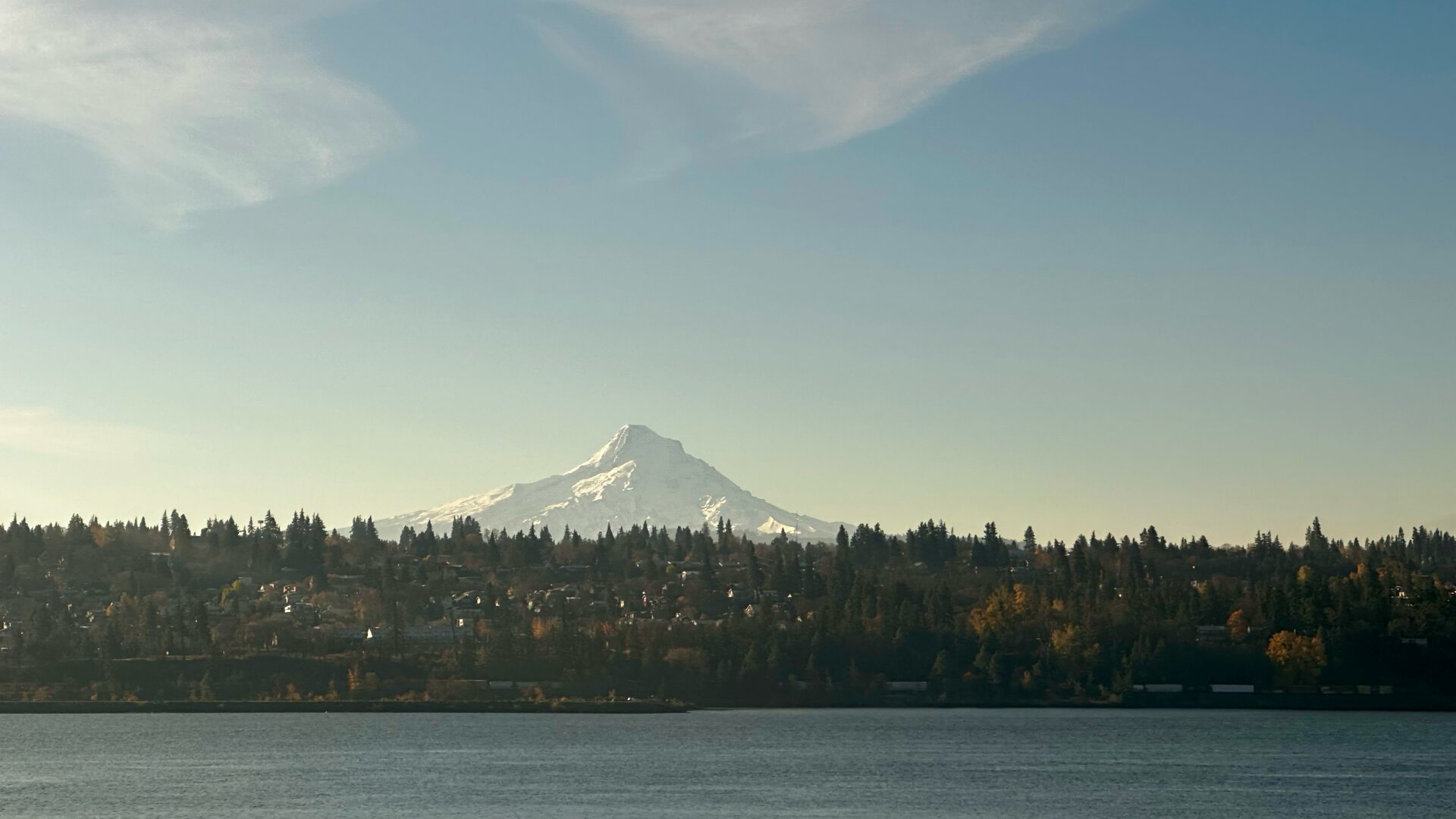 A pyramid shaped snow covered volcano rises above a city site on a hill rising from a river. The sky is mostly clear with some cirrus above. The mountain is lit from the left.