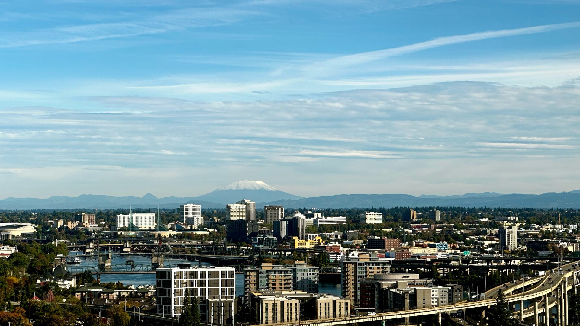 A snowcapped volcano rises in the horizon behind an urban setting with high rises, bridges, and a river.