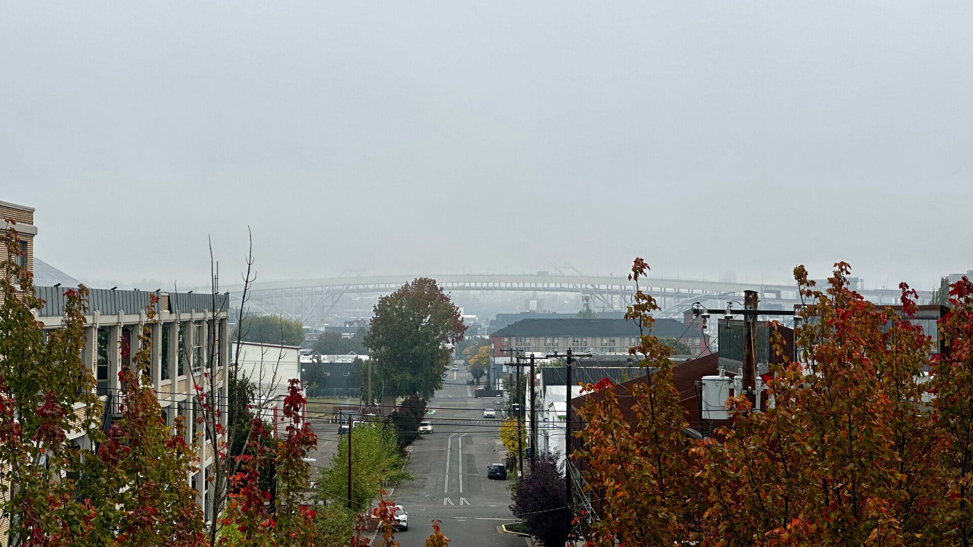 An urban industrial street scene with a bridge partially obscured by low clouds in the distance. Trees with fall colors are in the foreground.