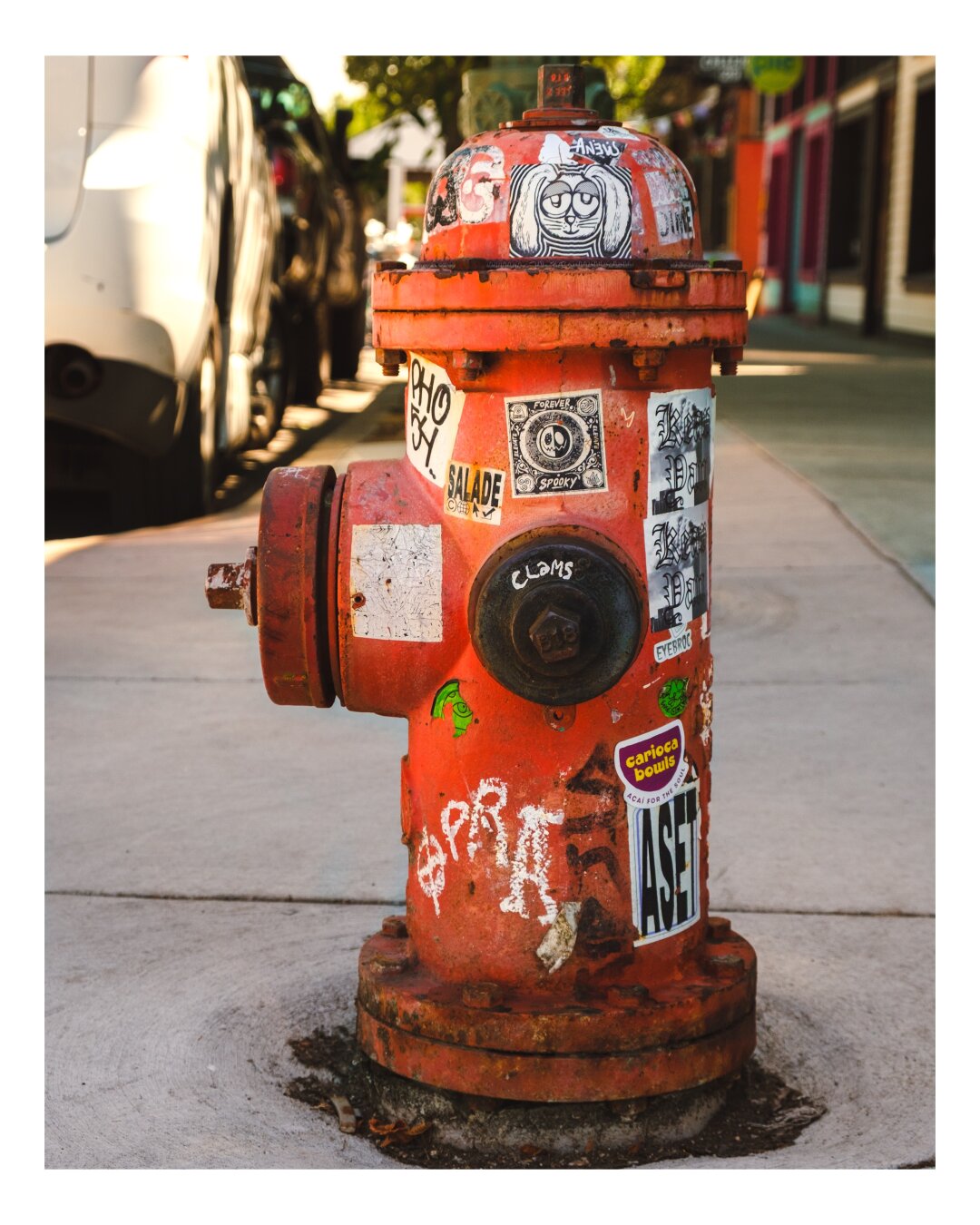 A weathered orange fire hydrant covered in stickers and graffiti stands on a sunny urban sidewalk with parked vehicles blurred in the background.