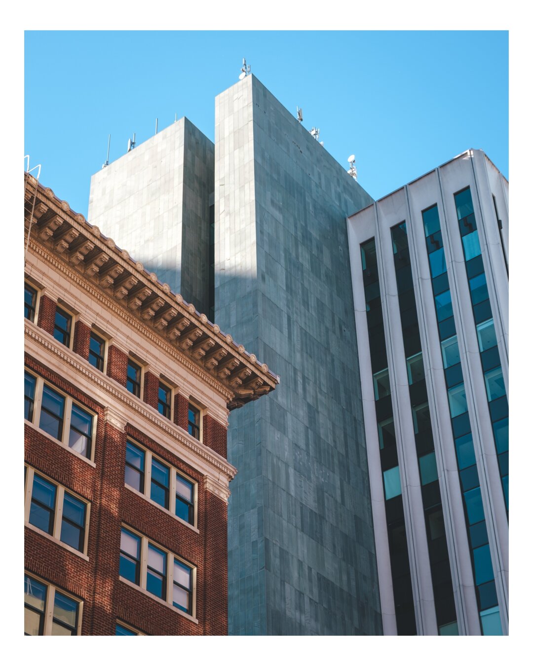 A warm brick structure, a stark concrete tower, and a modern glass facade converge under a brilliant blue sky.