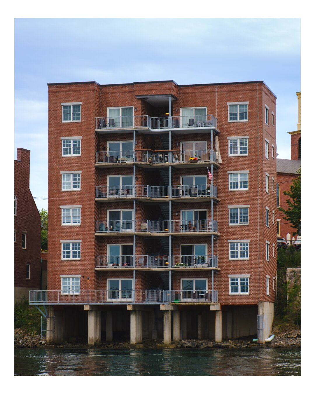A six-story brick waterfront apartment building with metal balconies and large windows, elevated on concrete pilings above the water, under an overcast sky.