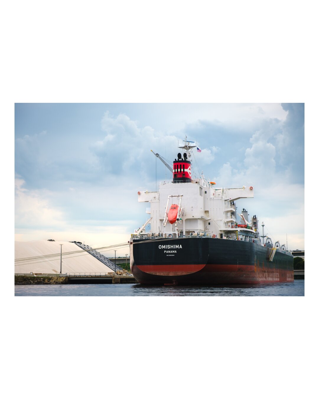 A massive industrial tanker ship with a red hull and white superstructure docked at a riverside terminal under a partly cloudy sky.