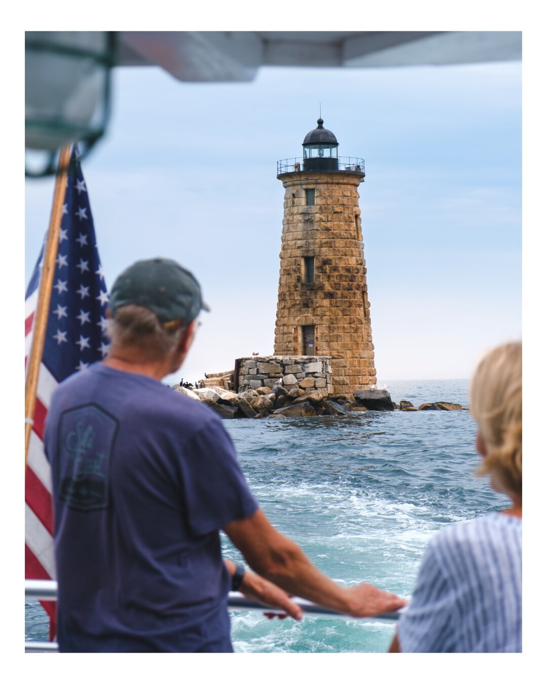 Two people aboard a boat gaze toward a solitary stone lighthouse standing on rocky waters under a serene sky, with an American flag visible in the foreground.