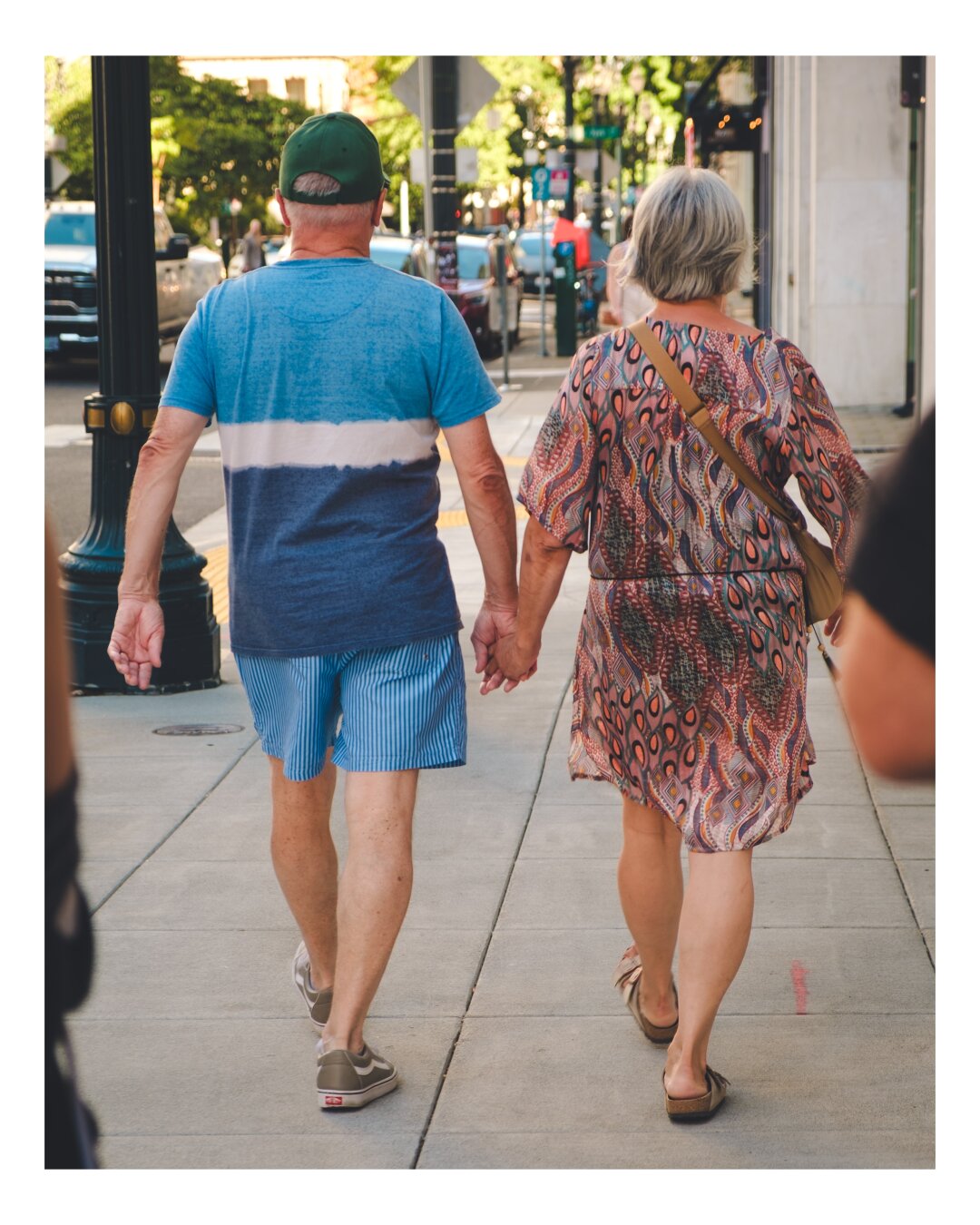 Two people holding hands in an walk along a sunny urban sidewalk lined with storefronts and trees.