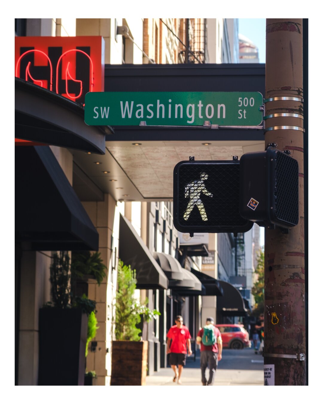 A pedestrian walk signal glows beneath a green street sign for SW Washington 500 on a sunny urban street lined with storefronts and tall buildings.