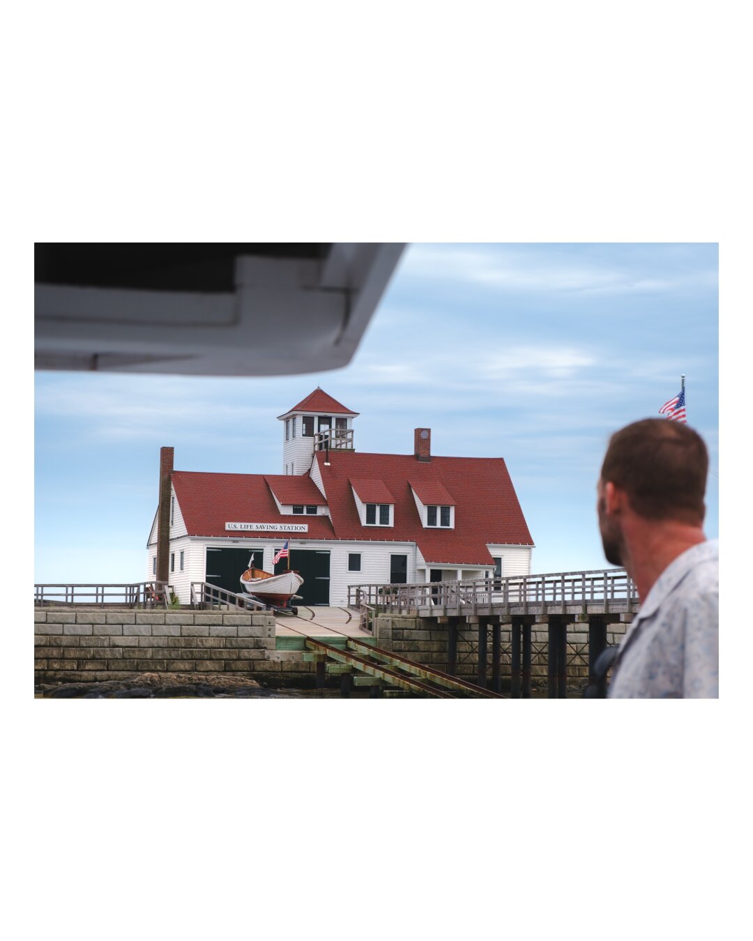 A historic red-roofed life saving station with white walls and a central tower stands on a stone pier, viewed from a boat on calm waters under a partly cloudy sky.