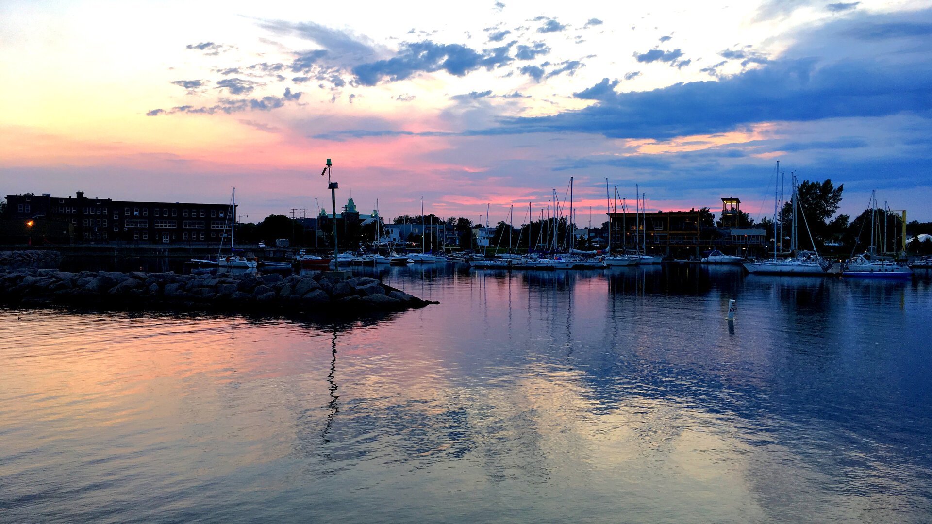 Photo of a marina with a few boats floating. Its sunset with a few clouds in the sky and many pink and purple colours reflecting in the water.