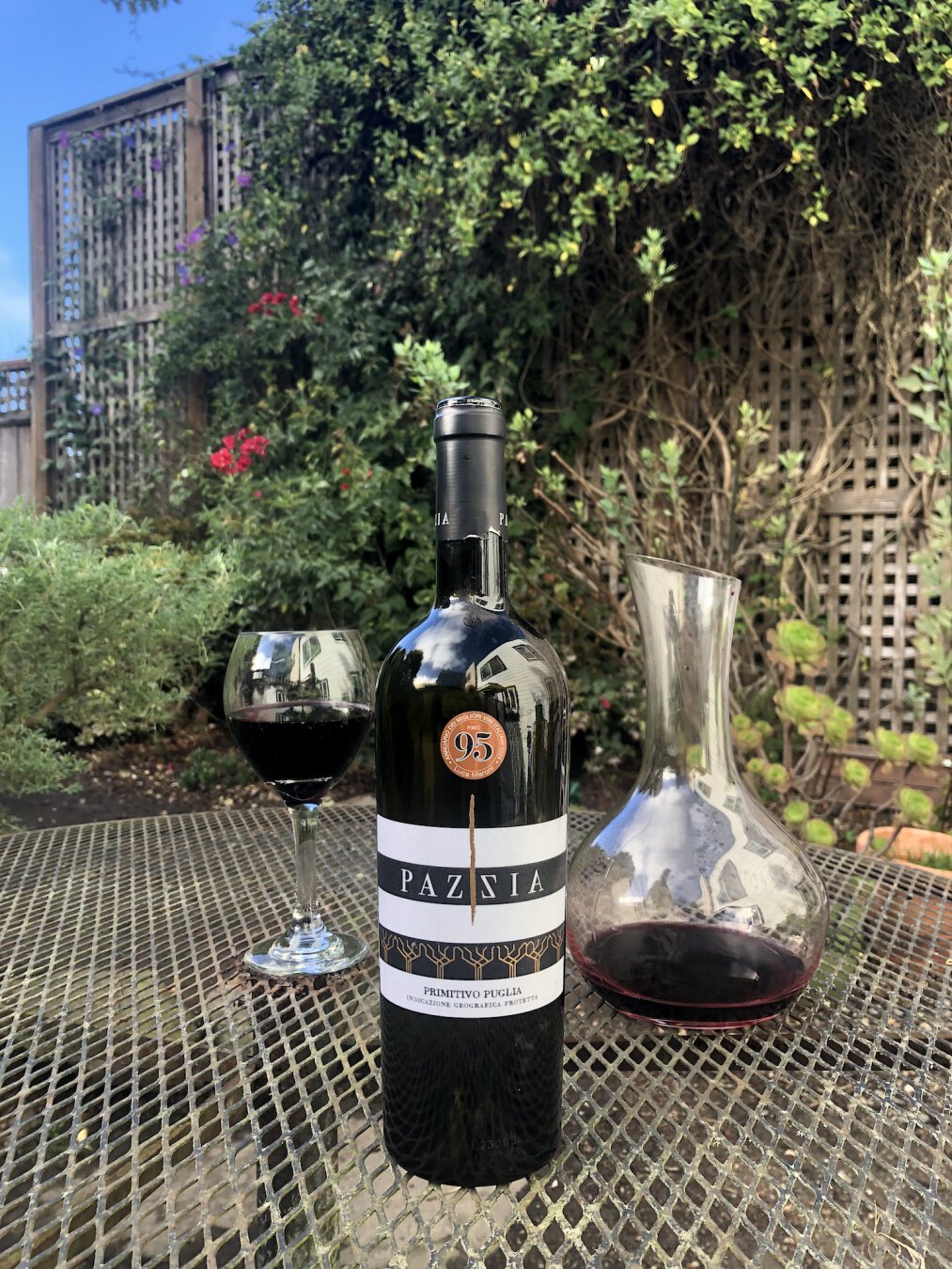 A dark wine bottle on a faded wrought iron patio table. Behind the bottle to the left, a glass with a full pour of red wine. To the right, a decanter of wine, mostly empty. In the background a large trellis in a garden with many vine growing on it. Toward the left end of the trellis, blue flowers climb to the top.  Beyond it, the sky is blue.

The bottle's label is a horizonally striped black and white. In the middle toward the top, the name PAZZIA. Near the bottom in smaller text, Primitivo Puglia. Below that in small text, Indicazione Geografica Protetta. In the upper third of the bottle, a round sticker displaying the number 95.