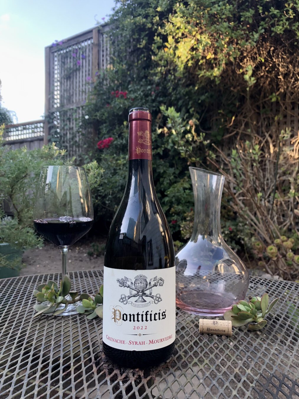 A bottle of red wine on an outdoor patio table. In the background to the left, a full pour in a large wine glass, surrounded by a sprig of jade. To the right, an empty decanter, woohoo! In front of that lies the cork, next to a sprig of jade.

Front and center, the wine bottle. The glass is dark in this light and is reflecting the house out of frame to the right. The label is off-white and features a coat of arms in the upper half. In the middle, the text Pontificis. Under that, 2022. At the bottom, Grenache - Syrah - Mourvèdre.

In the background, many green leafy plants and a tall, wide trellis which is full of leafy vines.