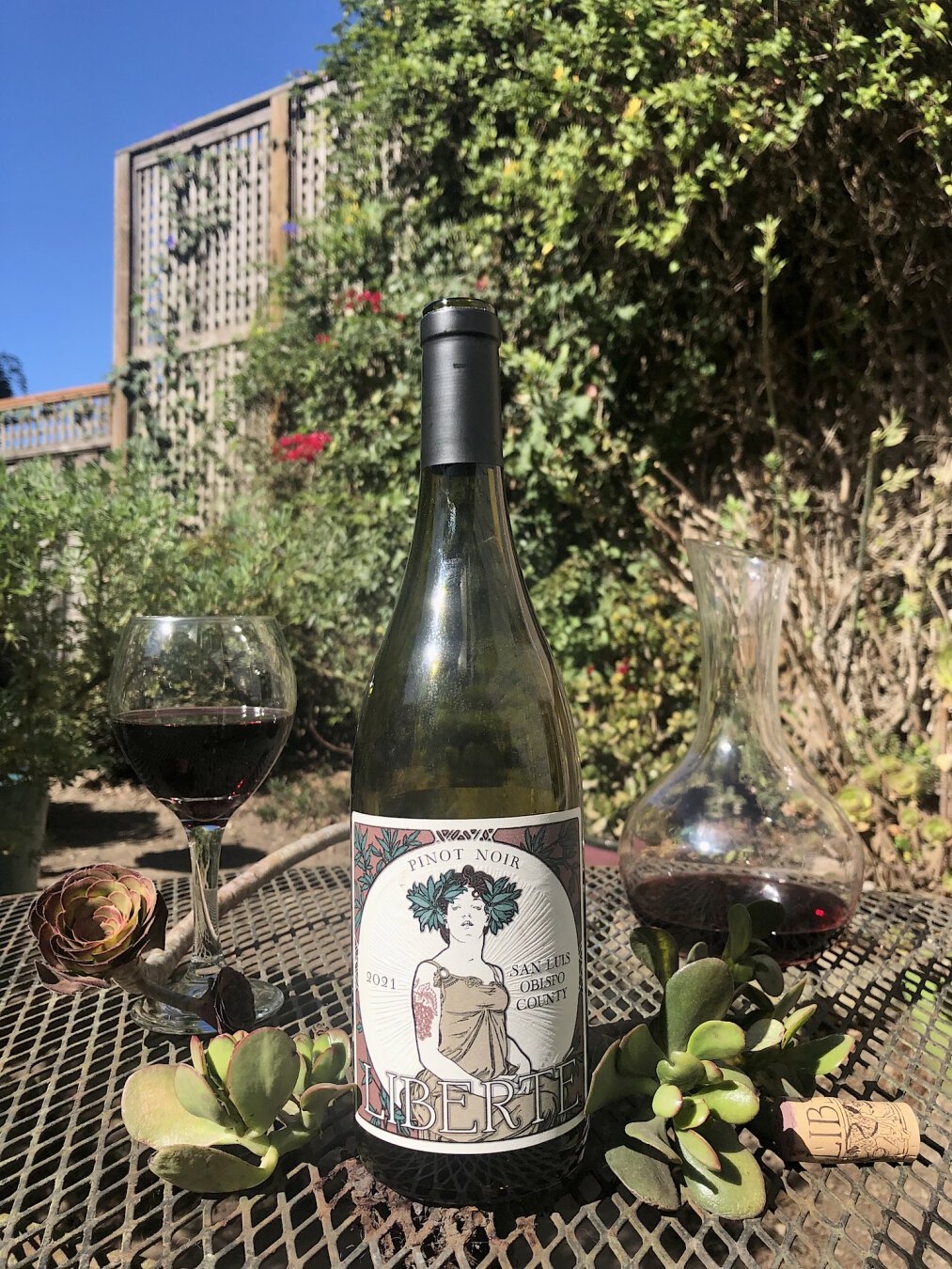 A bottle of red wine on a black wrought-iron patio table, outdoors. To either side, sprigs of jade and to the right, the cork. A glass of red wine is in the near background to the left and a decanter of wine is in the background to the right. A succulent drapes itself around the glass. In the background and out of focus, a trellis full of vines illuminated by the Sun. The sky is a bright blue.

The off-white and green embossed label sports a drawing of a woman which owes much of it's style to the ancient greeks. Her hair has flowers in it and she's wearing a sleeveless dress. A large tattoo depicting grapes is visible on an upper arm.

At the top middle, the text Pinot Noir. To the left of the woman, 2021. To the right, San Luis Obispo County. At the bottom in large stylized font, Liberté.