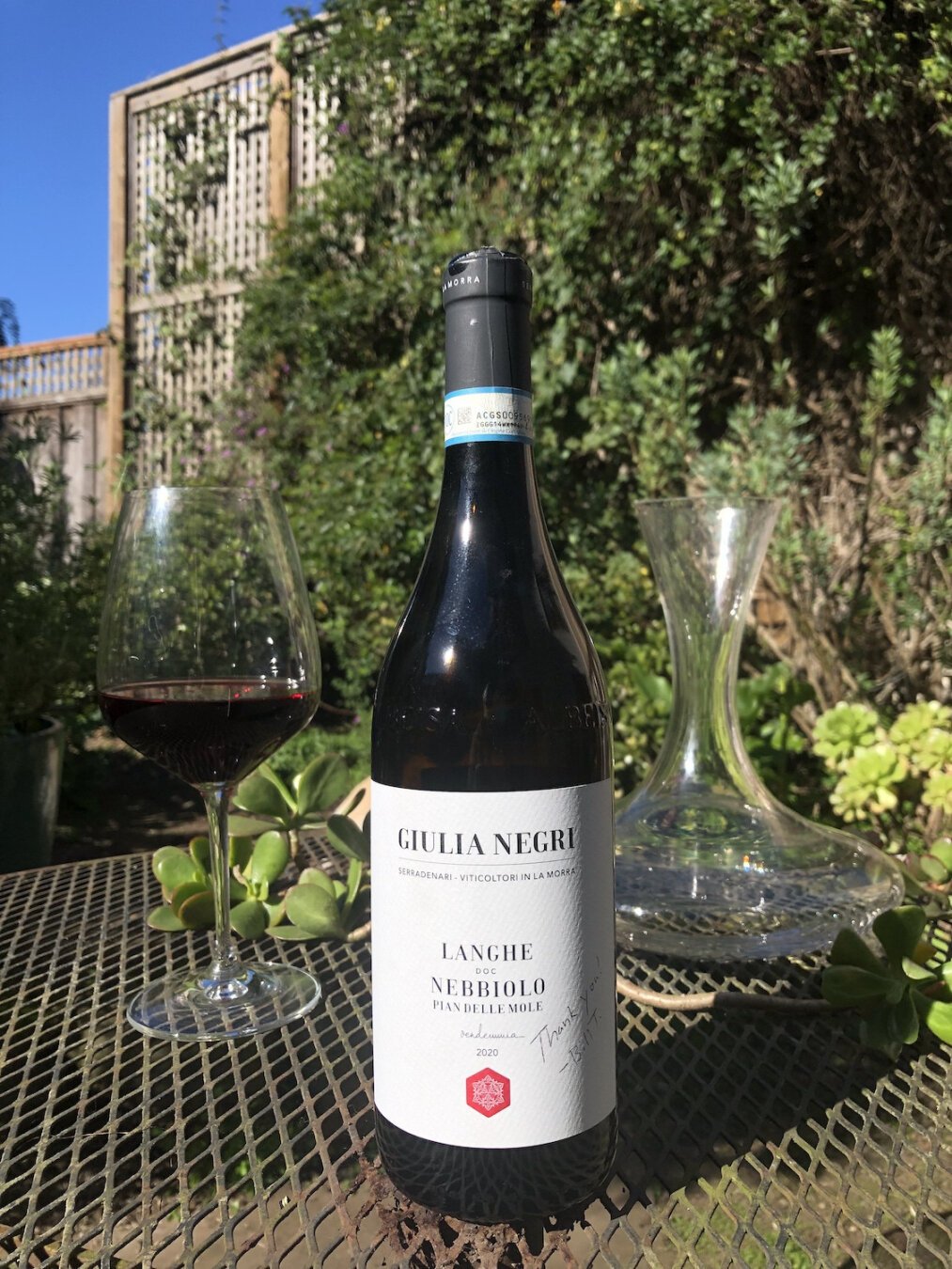 A dark bottle of wine on a garden table. A glass of red wine is in the background on the left, and an empty decanter is to the right. There are many green plants out of focus in the background, and a wooden trellis can be seen in the background next to a brilliantly blue sky. The bottle's label is lit up by sunshine, and is white with black text. At the top, it reads Giulia Negri. Next line, Serradenari Viticoltori In La Morra. In the center of the label is the text, Langhe DOC Nebbiolo. Next line, Pian Delle Mole. At the bottom, the year 2020.