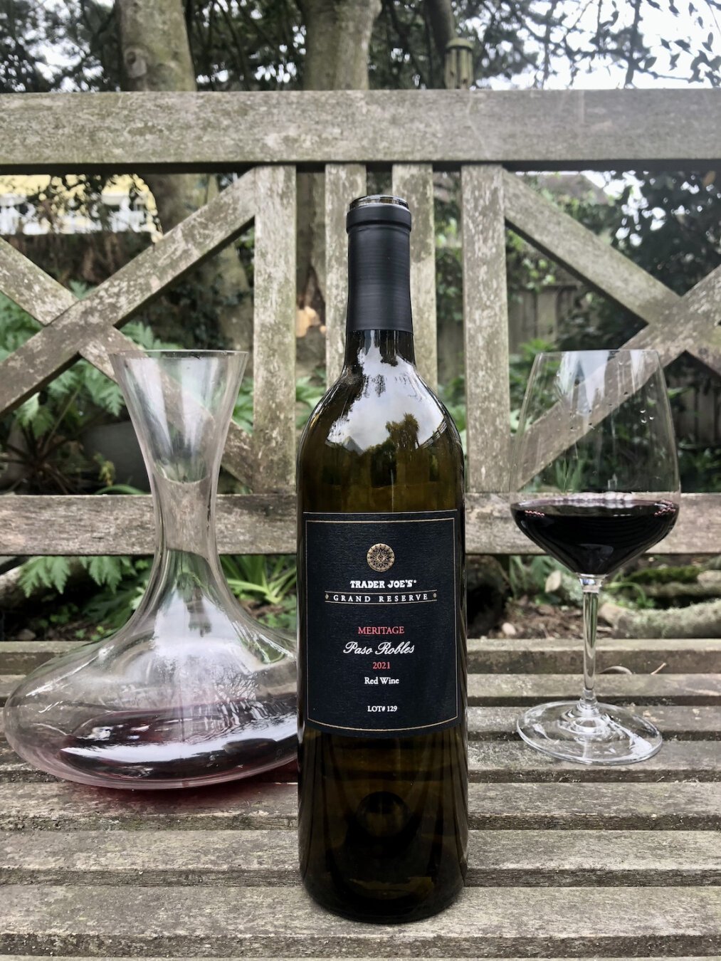 A semi-transparent empty wine bottle on a weathered garden bench. To the left in the background, a decanter of red wine. To the right, a glass with a full pour of red wine. In the background, out of focus plants and trees. 

The bottle's label has a dark background with white and gold text. At top, a round decorative symbol. Under that, Trader Joe's Grand Reserve.  At vertical center, Meritage Paso Robles 2021 Red Wine. At bottom, Lot #129.

The sky, miscellaneous plants and a house are reflected off the top of the bottle.