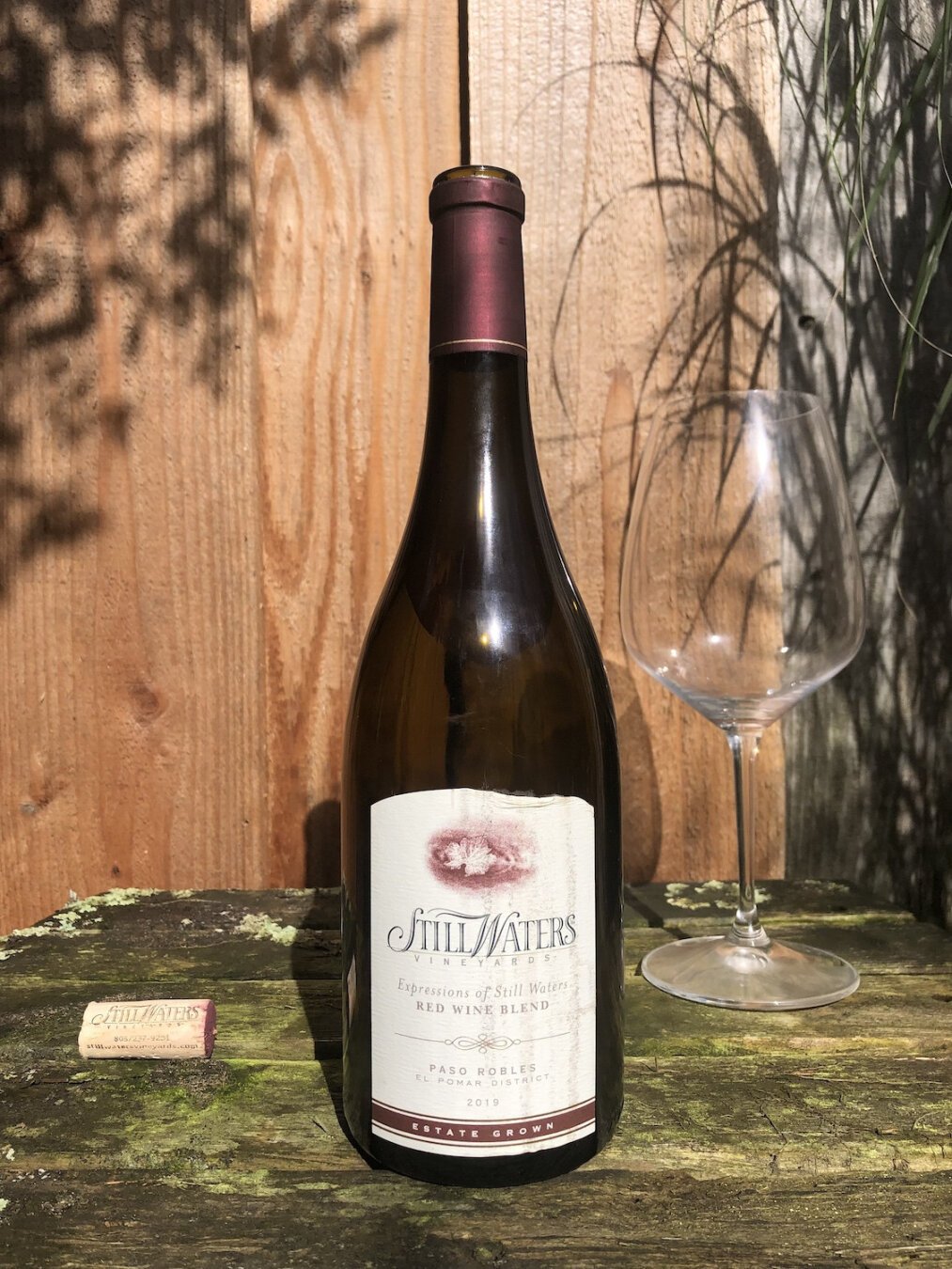 A bottle of red wine on a distressed wooden table. An empty wine glass is in the background to the right, and a cork with the text Still Waters is to the left. The bottle is lit by sunlight, as is a wooden fence in the near background. Shadows of plants can be seen on the fence.

The bottle label is white with dark text. An artsy image of a vine leaf is at top center. Below that in a large font, Still Waters Vineyards. Below that, Expressions of Still Waters Red Wine Blend. At bottom, Paso Robles El Pomar District 2019 Estate Grown.