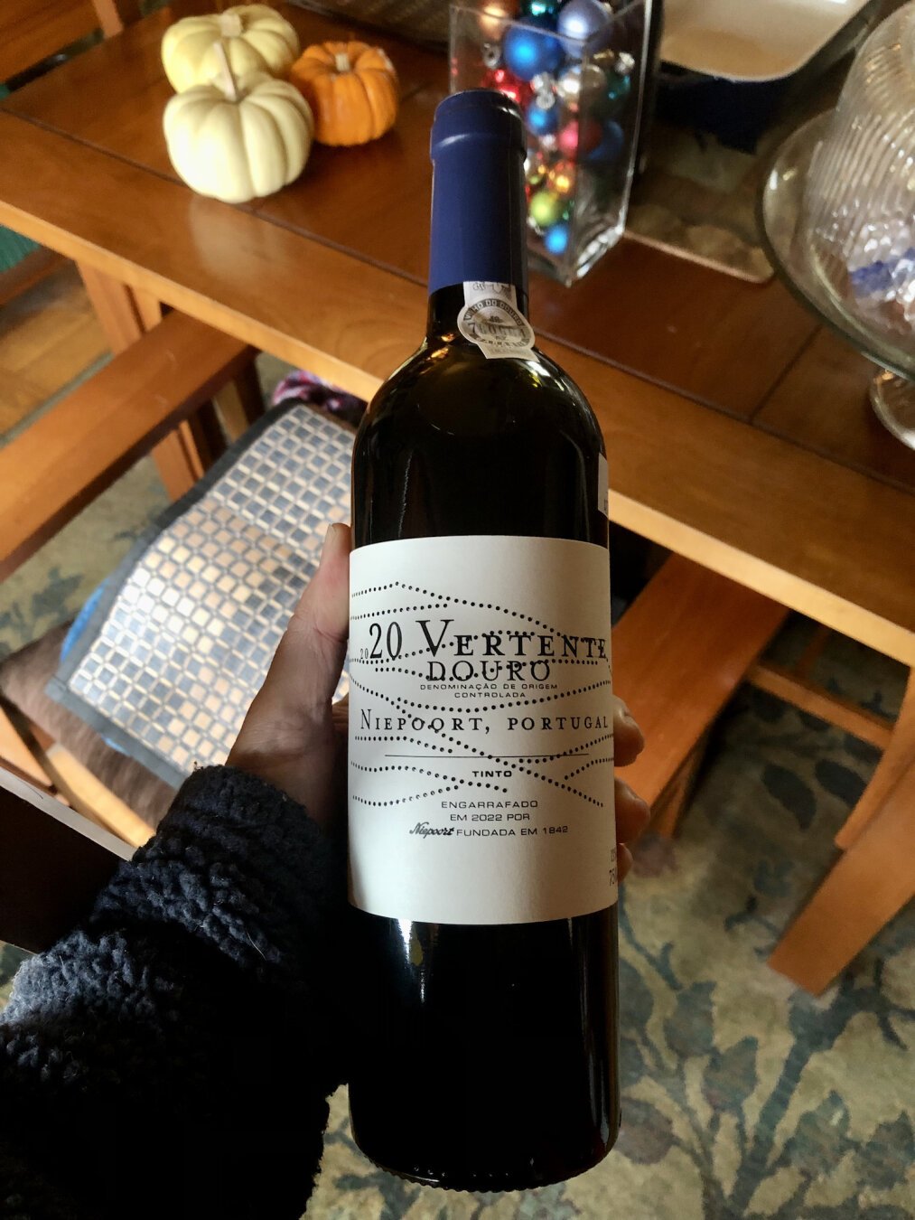 A closeup photo of a bottle of red wine, being held above a cherry dining room table. The glass bottle is dark, and the label is white with black text. Interspersed amongst the text are dotted black lines both horiztonal and diagonal, which suggest hills to me. 

At top, the text reads 2020 Vertente Douro. Next lines, Denominação de Origem Controlada. 

Below that, Niepoort, Portugal, followed by Tinto.

Near the bottom, Engarrafado Em 2022 Por Niepoort Fundada Em 1842.