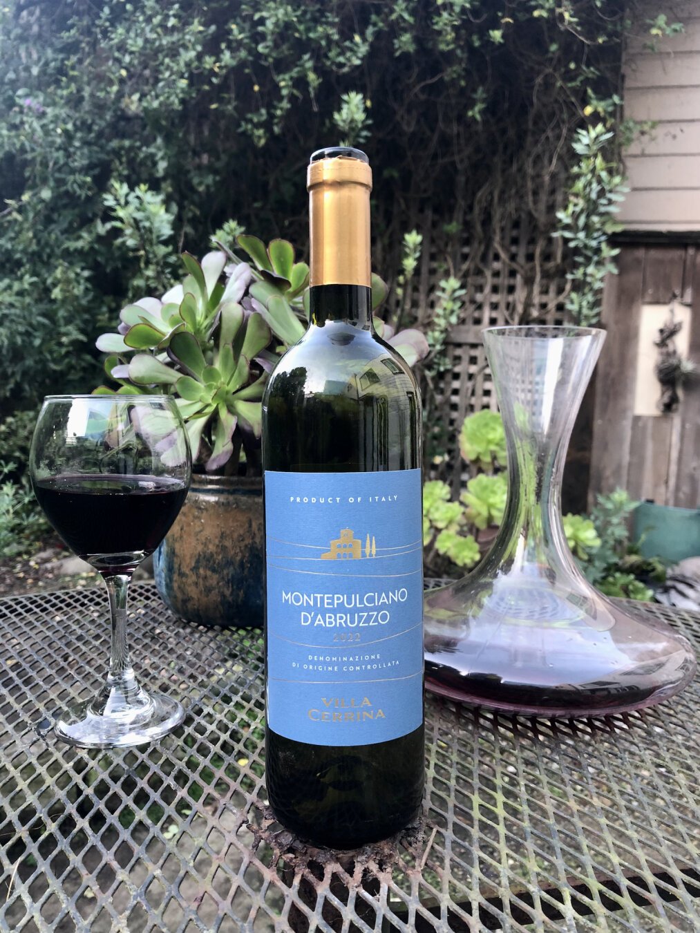 A bottle of red wine on an outdoor patio table. The bottle has a light blue label with wine text and gold accents. At the top of the label is the text Product of Italy. A gold image of a villa comes next, with two Italian Cypress trees to the right. Center of the label are the words Montepulciano d'Abruzzo. Below that, 2022. Next Denominazione di Origine Controllata. At the bottom, Villa Cerrina. 

To the right of the bottle in the background, a decanter with some red wine. To the left of the bottle in the background, a glass of wine. In the distance can be seen various garden plants and a trellis.