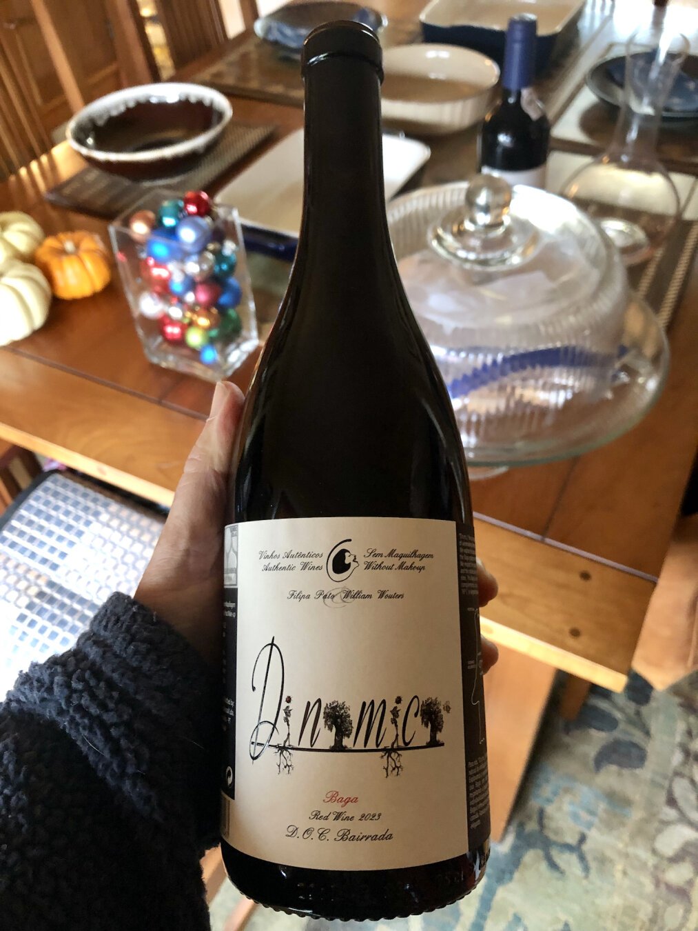 A closeup picture of a bottle of red wine. It's a dark bottle being held above a cherry dining room table with a full place setting. 

The label is soft white with black writing. At the top in both Portuguese and English is the text Authentic Wines Without Makeup. Beneath that, the names of the principals, Filipa Pato - William Wouters.

The name of the wine, Dinamica is in the center and printed in a playful typeface, with four of the eight letters being replaced by drawings of vines, with pollinators hovering around them.

At bottom is the text Baga, followed by Red Wine 2023. At bottom, D.O.C. Bairrada.