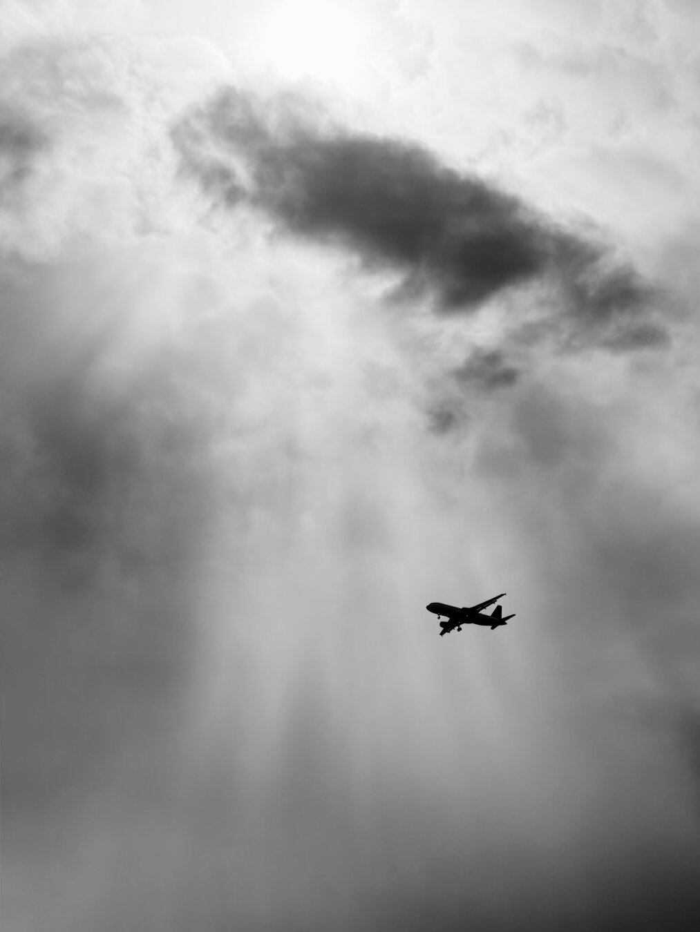 An Airbus A320 operated by Delta Airlines as DAL850 from Atlanta to DCA is seen under a cloudy sky with rays of sun attempting to shine through. The picture is in black and white and the plane has tail number N326US which can’t be seen from this distance.