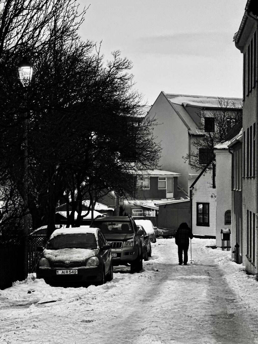 Black and white picture of a man with a cane walking down a snowy street. There are cars parked on the left side.