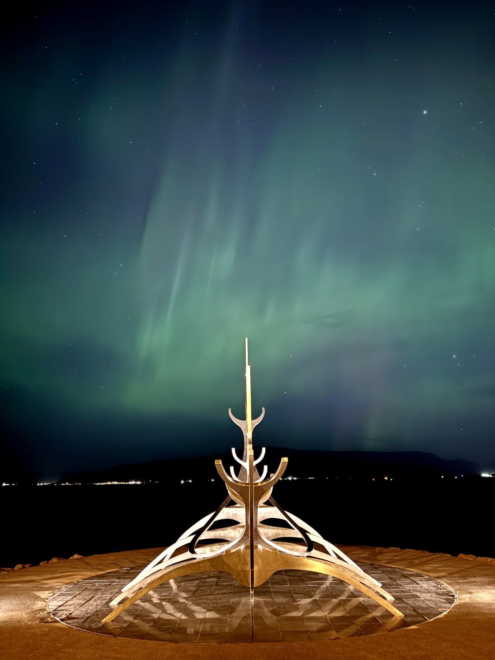 Sólfar, a sculpture of a Viking ship on the shore of Reykjavík, is lit up at night. The sky is also lit up with northern lights in green.