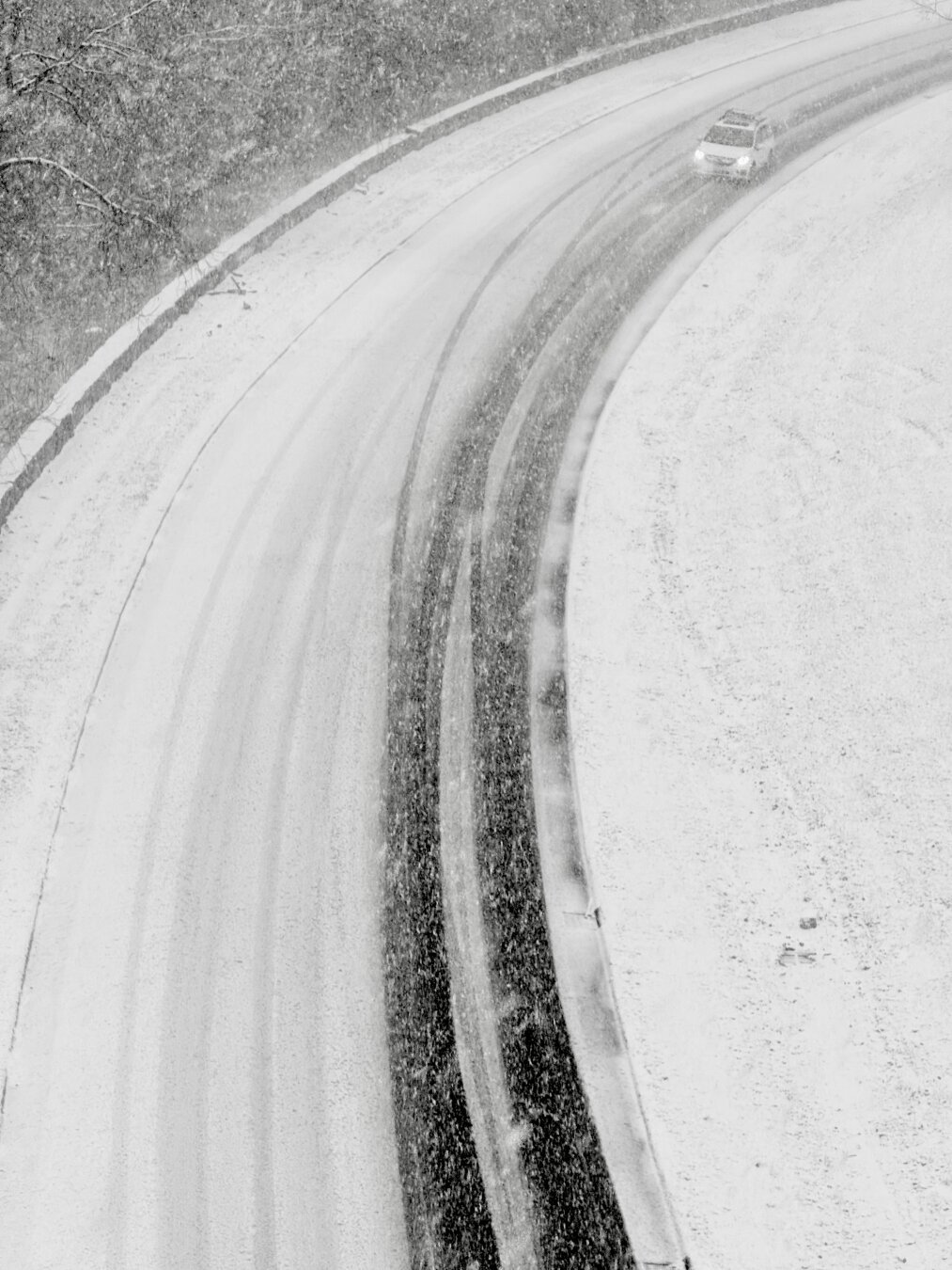 The GW Parkway is seen from the Key Bridge. The roadway is mostly covered in snow except for two tracks where car tires would go. There’s one car at the top right of the image making its way around the corner.