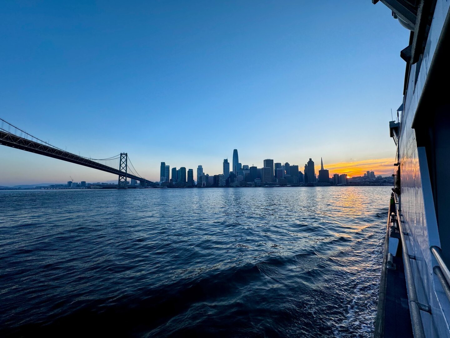 Wide shot of the Bay Bridge on the left, the city of San Francisco, the sunset, and the ferry on the right