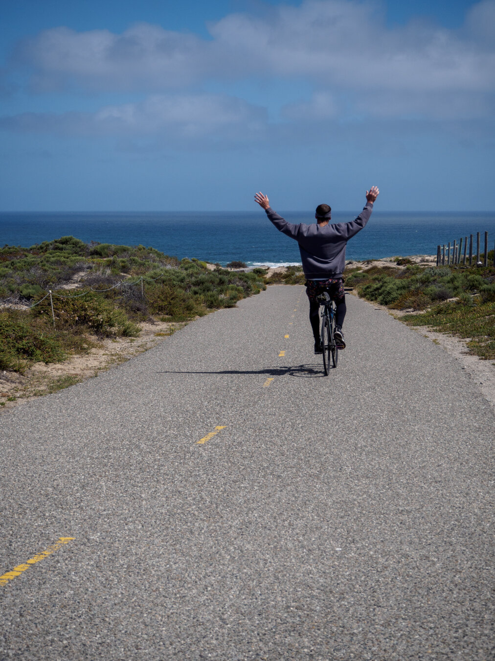 A cyclist riding down a paved hill towards the beach with his arms up in the air
