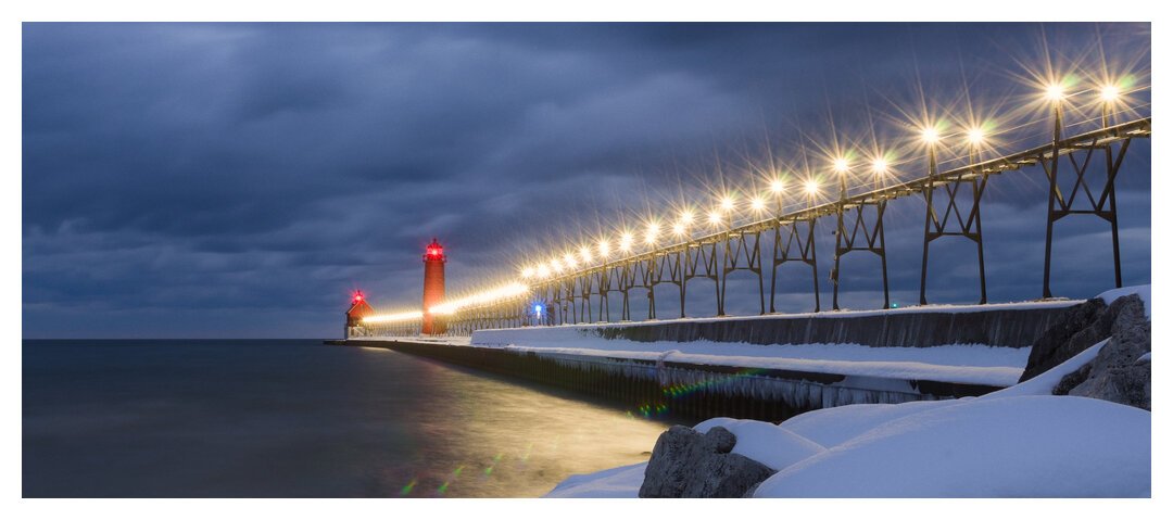 A wide view of a long concrete pier jutting out into Lake Michigan with a catwalk with lights on it along the pier. At the end are a red lighthouse and secondary building with red lights on top. A moody, cloudy sky looms overhead.