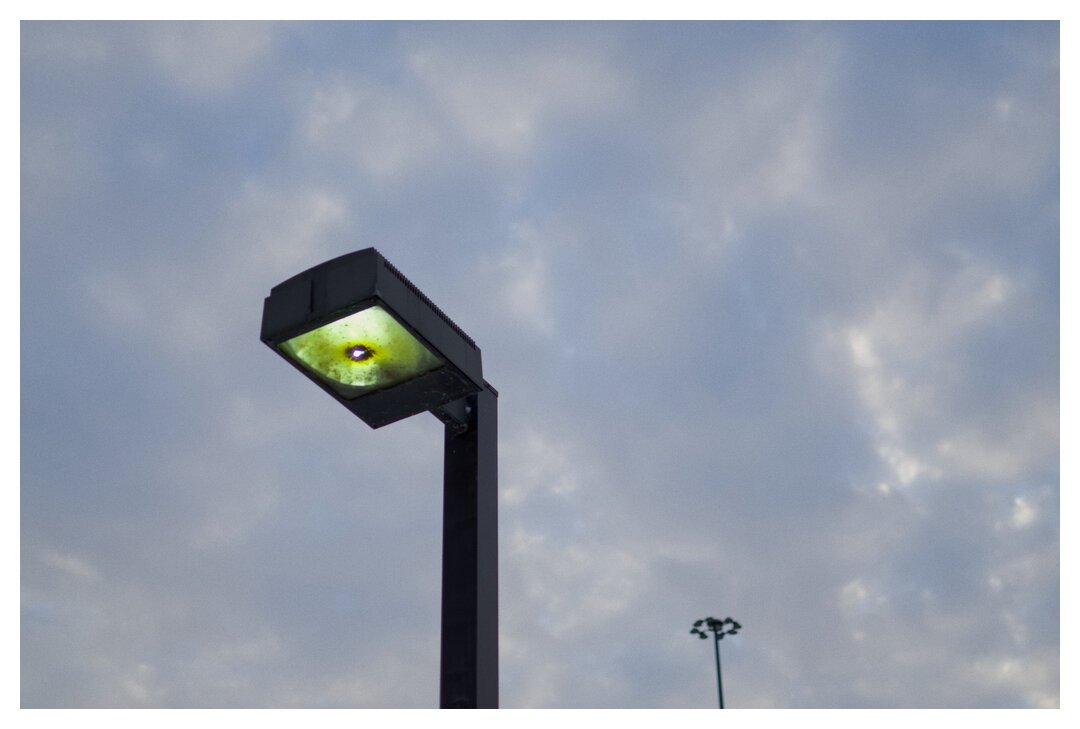 A street lamp with a greenish-yellowish hue with a larger street lamp with many lights on it in a circle in the background. Set against a cloudy sky.