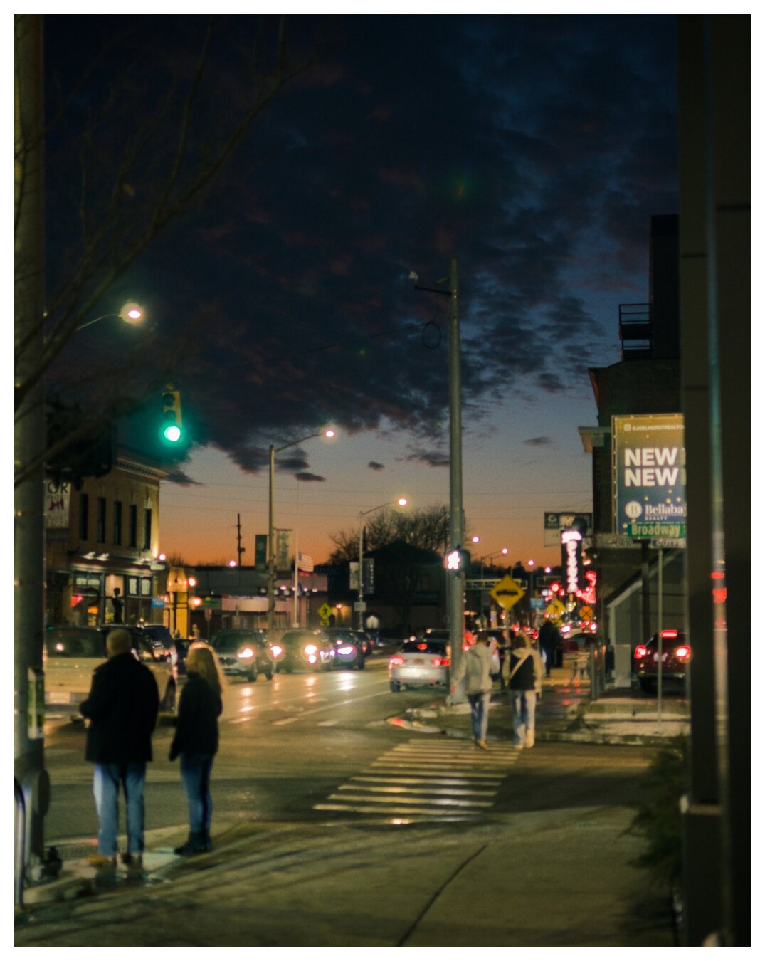 A city intersection with a few cars and a cloudy, dark sunset in the background. A few people linger on one corner while a few others are crossing the road on a crosswalk.