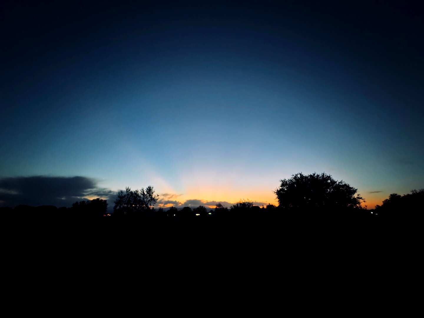 Sunset over Colorado’s foothills. Trees contrast a dramatic orange-blue gradient and faint sun rays in the background.