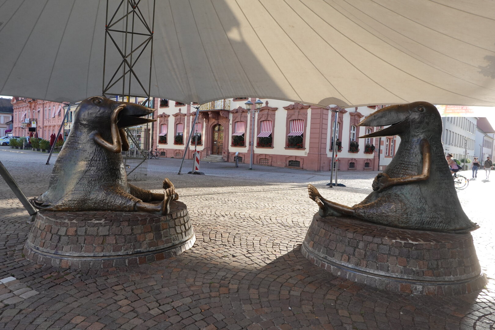 Two giant bronze raven sculptures, sitting like people with their legs stretched out, under a canopy in Offenburg's central square. In the background, a pink-and-white historic building with a balcony and windows adorned with red flower boxes.