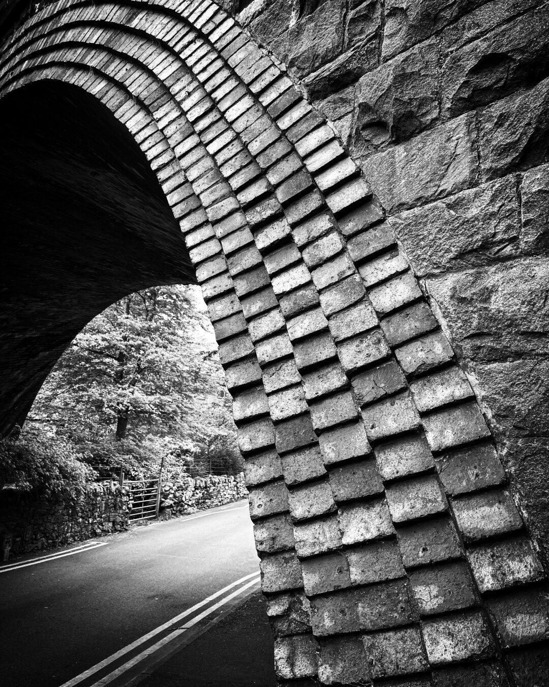 Close up of a railway bridge arch. The picture is in black and white and shows the detail of the rows of bricks. Also visible is the road running bottom left towards top right. The bridge is near Snowdon in Wales.