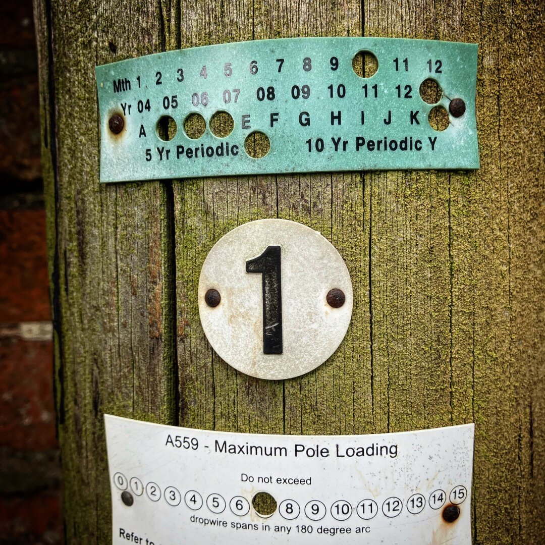 A photo of 3 identification labels on a UK telecoms pole. Focus is on the centre label that is a white circle with a black number one.