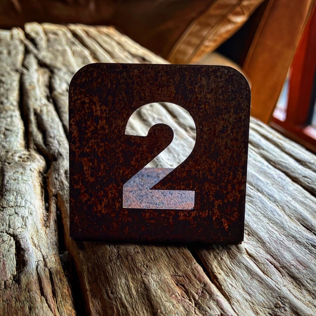 A metal restaurant table number. Laser cut from thin metal. It’s the number two. It is sitting on a very rustic wooden table.