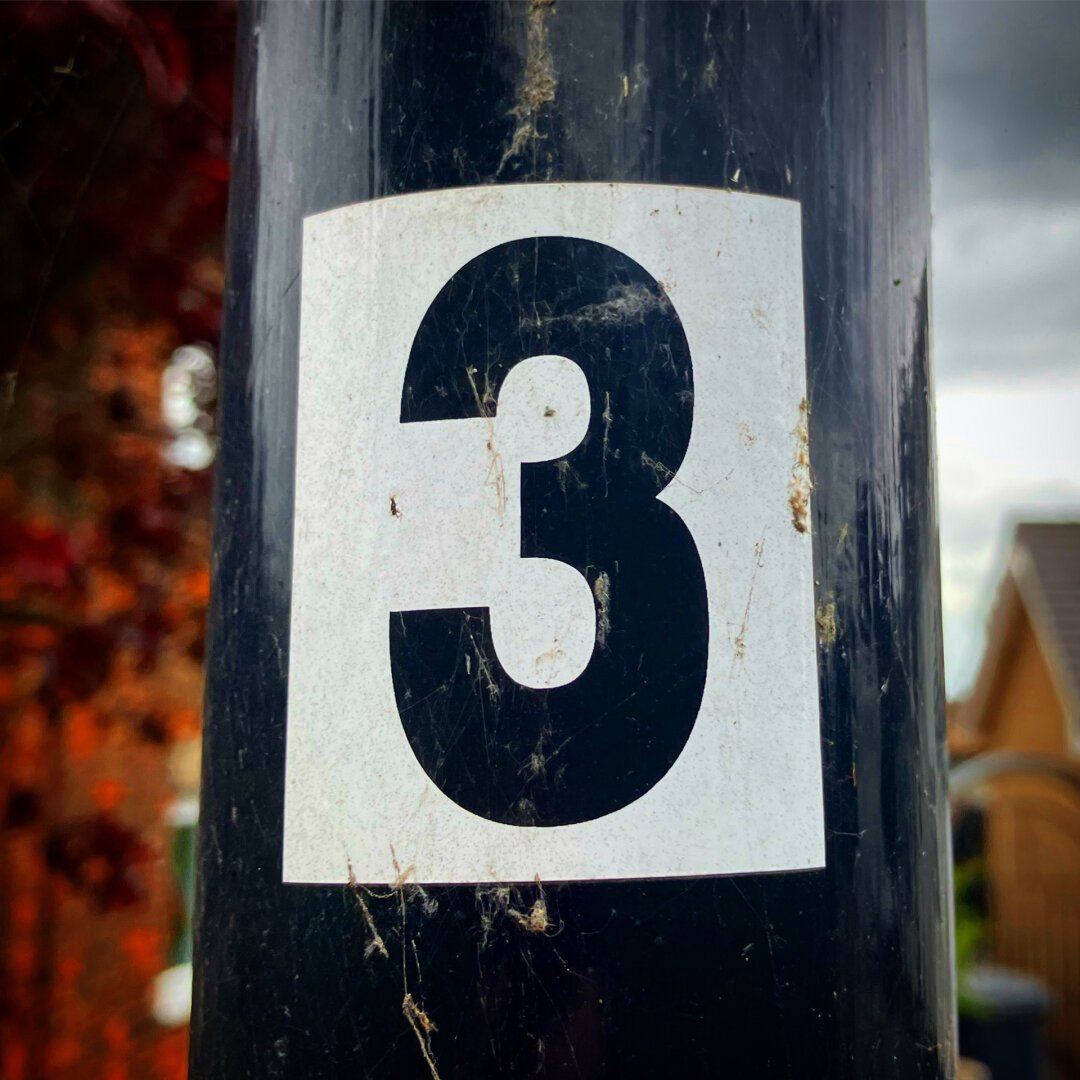 A square white vinyl sticker bearing the number three in black. It is on a black utility pole.